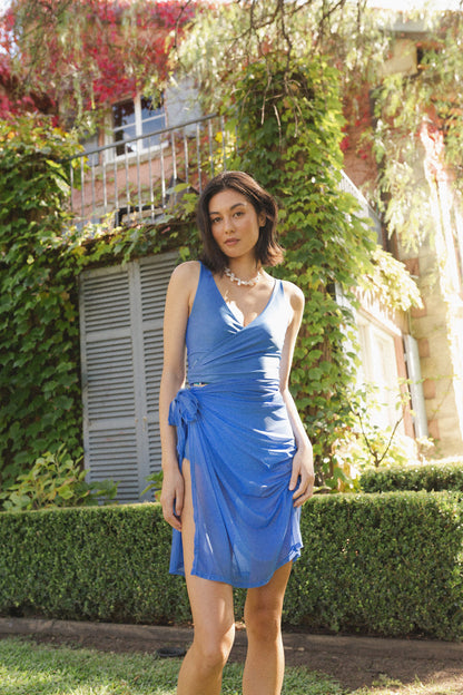 Woman stands in garden wearing a blue bikini set, with a matching sarong tied around her waist.