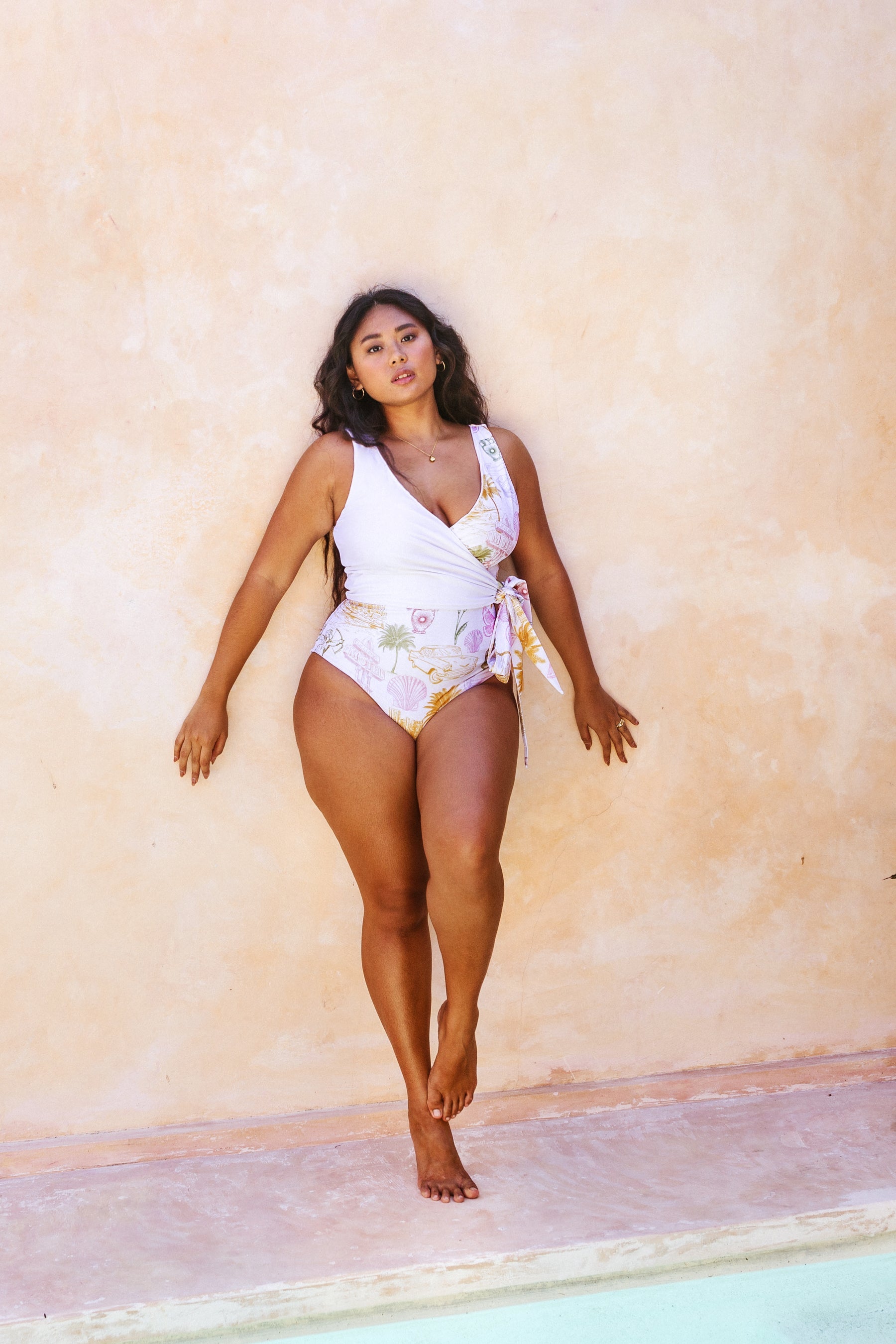 Woman leaning against a stone wall by the pool dressed in the colourful vintage post-card motifs printed the Havana white reversible wrapsuit with matching waist tie.