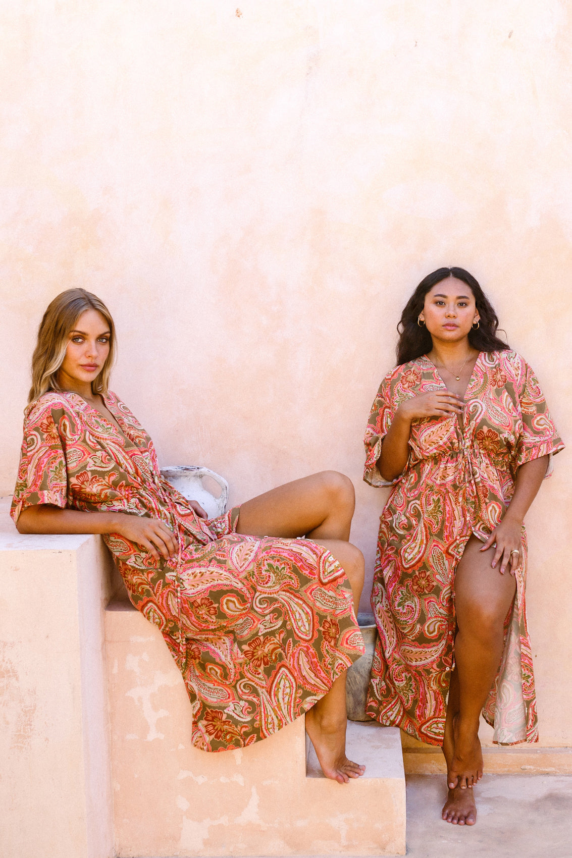 Two women posing, one sitting on a stone staircase and another standing against a stone wall, dressed in the Barcelona kaftan dress.