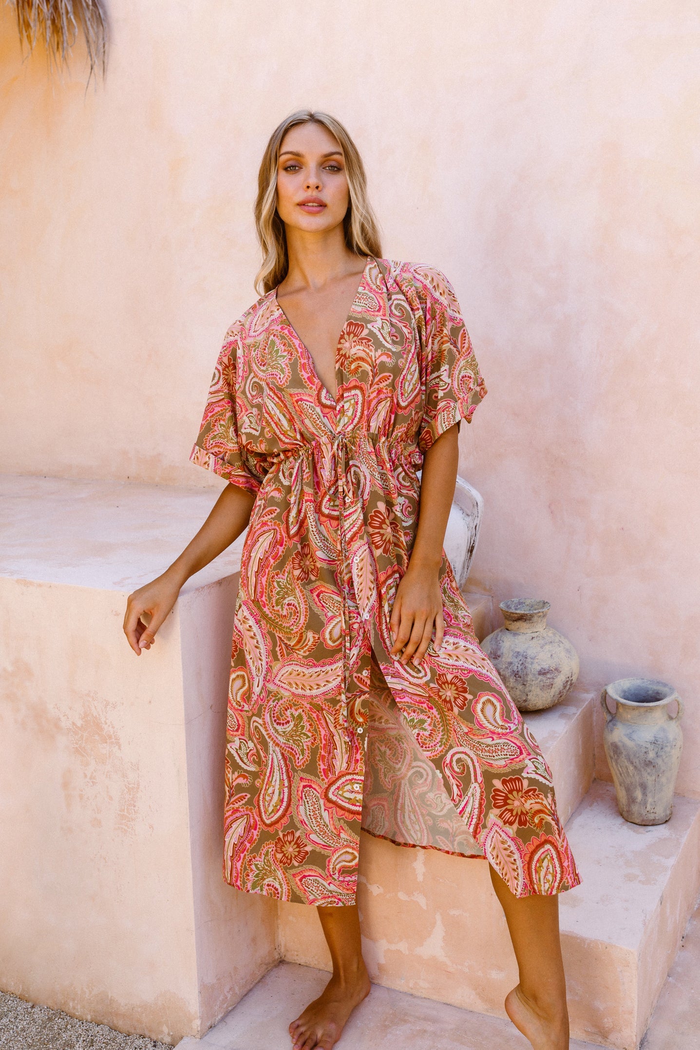 Woman posing in front of stone staircase and artisanal vases dressed in the paisley pink print kaftan dress