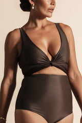 Woman stands in front of a beige background wearing a deep chocolate brown coloured bikini set. The top is cinched at the bust with a tie and the bottoms are high cut.