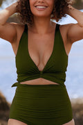A woman standing on a beach wearing a deep green bikini set smiles as she runs her hands through her hair. 