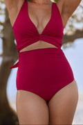 A woman wearing a vibrant raspberry pink coloured bikini set stands in front of a tree with her hands above her head. The bottoms are high waisted and the top is cinched at the bustline.