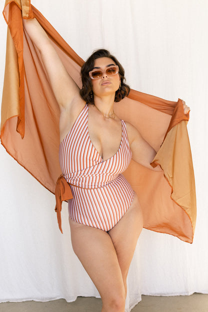 A woman wears a vertical striped orange, white and cream bikini set. She holds a matching rust coloured sarong aloft behind her.