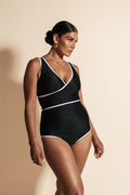 A woman stands against a beige backdrop wearing an all black bikini set with white binding.