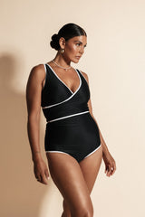 A woman stands against a beige backdrop wearing an all black bikini set with white binding.