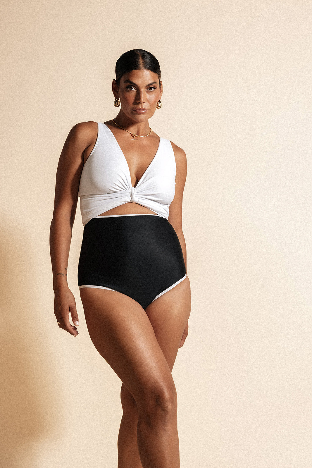 Woman standing in front of a beige wall wearing a white bikini top cinched at the bust, paired with high waisted all black bikini bottoms framed by white binding.