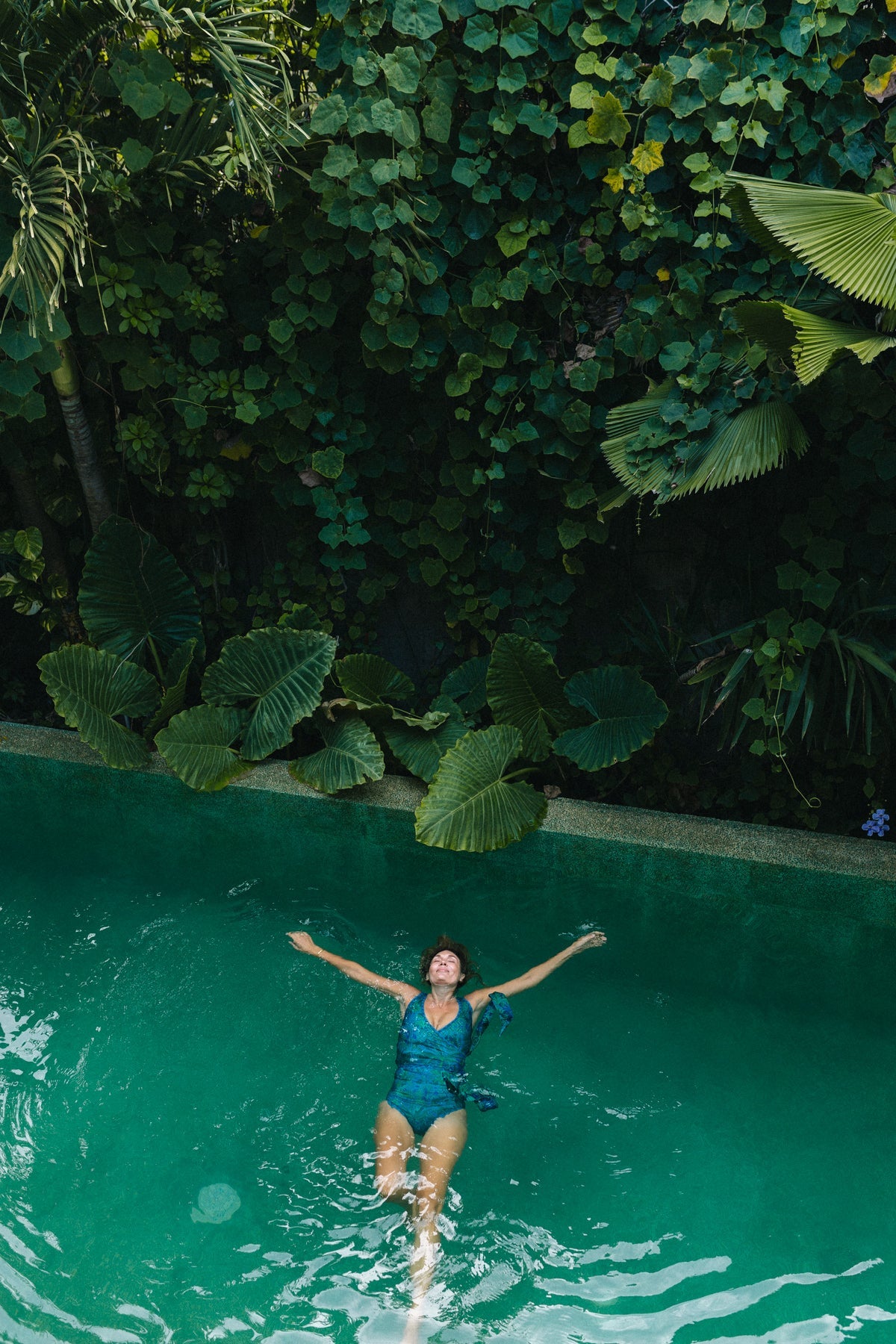 Woman floating on her back in a luxurious pool surrounded by lush green foliage, wearing a green and teal motif printed wrapsuit.